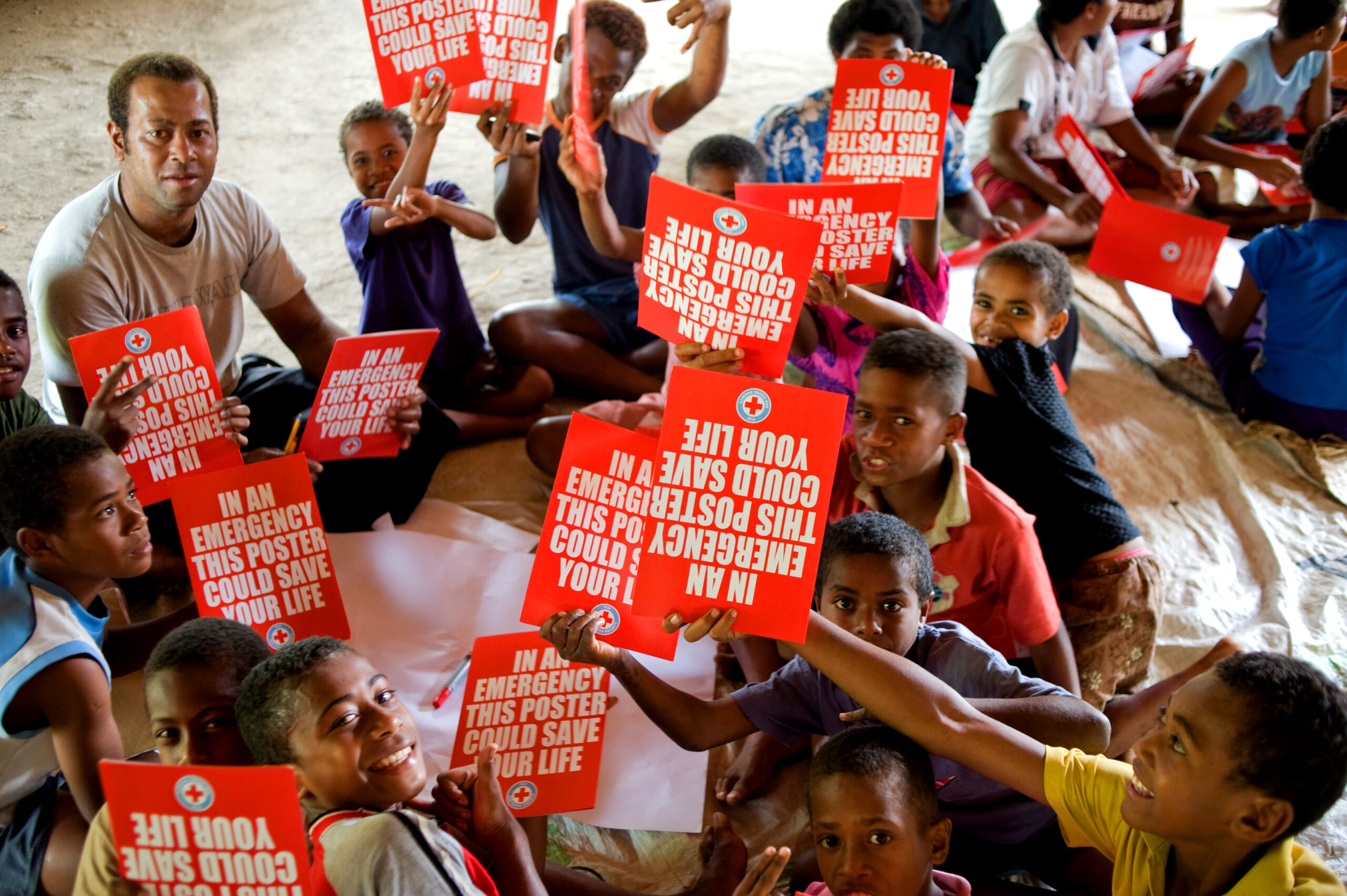 A large group of schoolchildren holding red posters with a Red Cross logo stating "In an emergency, this poster could save lives."