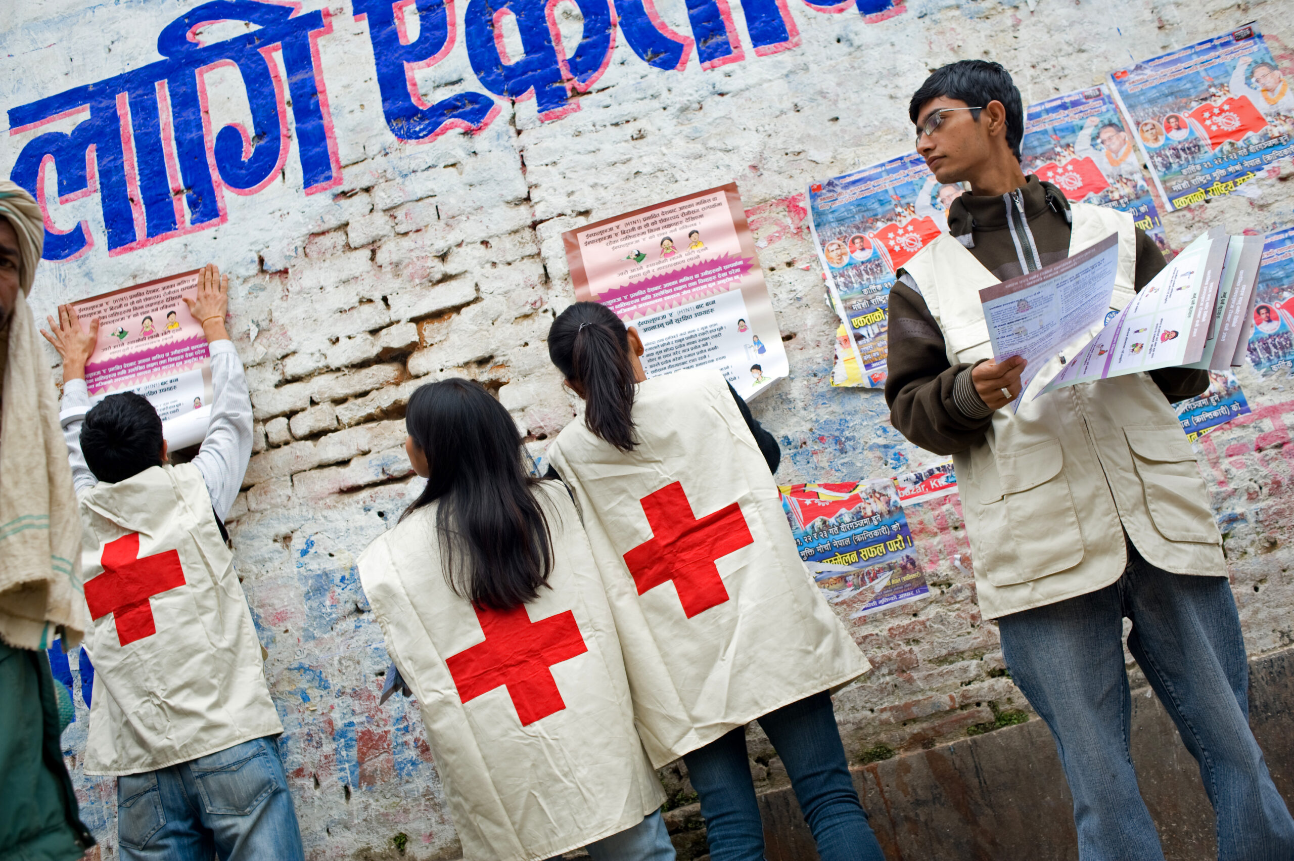Image shows a group of volunteers distributing pape materials in a street, they glued posters to walls for pedestrians to have access to public health information.