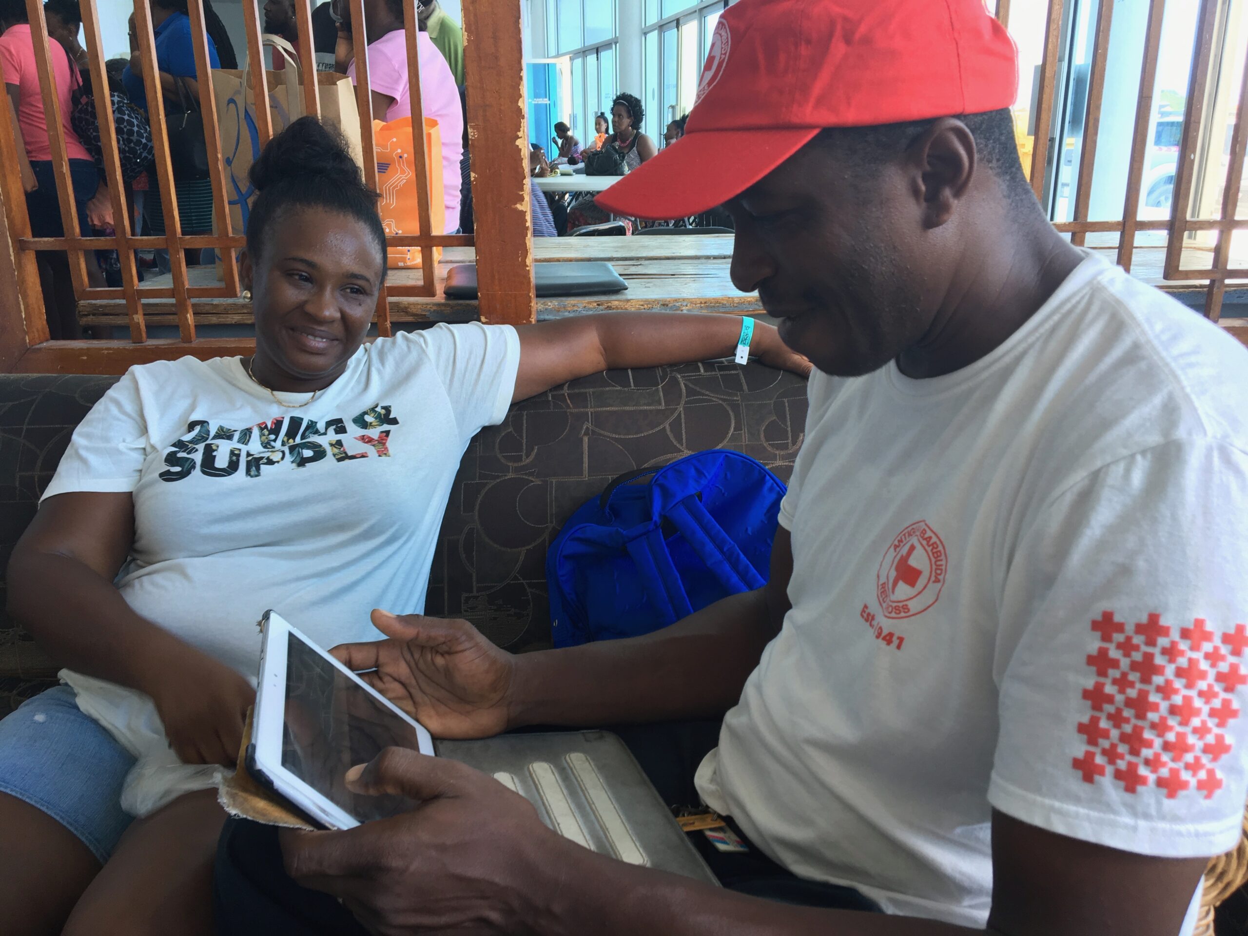 The image shows two smiling people reviewing documents on a tablet in an informal setting. One person wears a t-shirt with a graphic design, while the other has a red cap and a white shirt with a red cross logo.