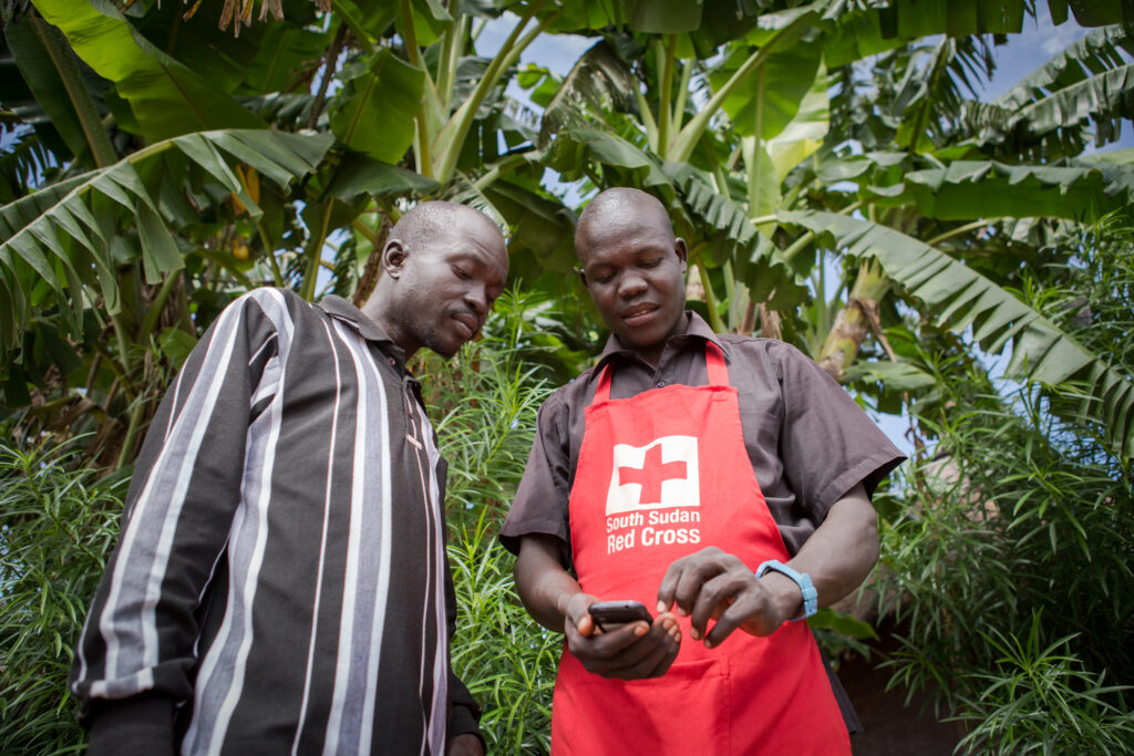David Luate, in red SSRC apron, handles a mobile phone enabled to assist the national government's Polio vaccination program. As a volunteer supervisor, Luate trains field volunteers to effectively use the technology.