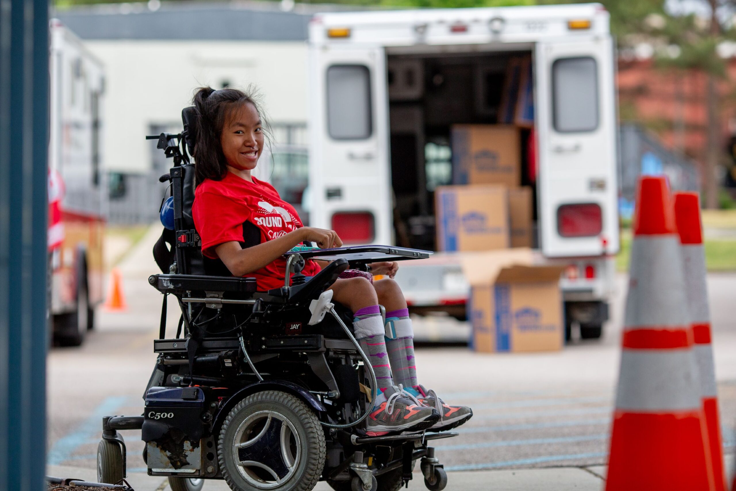 A smiling young girl in a wheelchair, wearing a red t-shirt, next to an emergency vehicle with open doors and supplies.