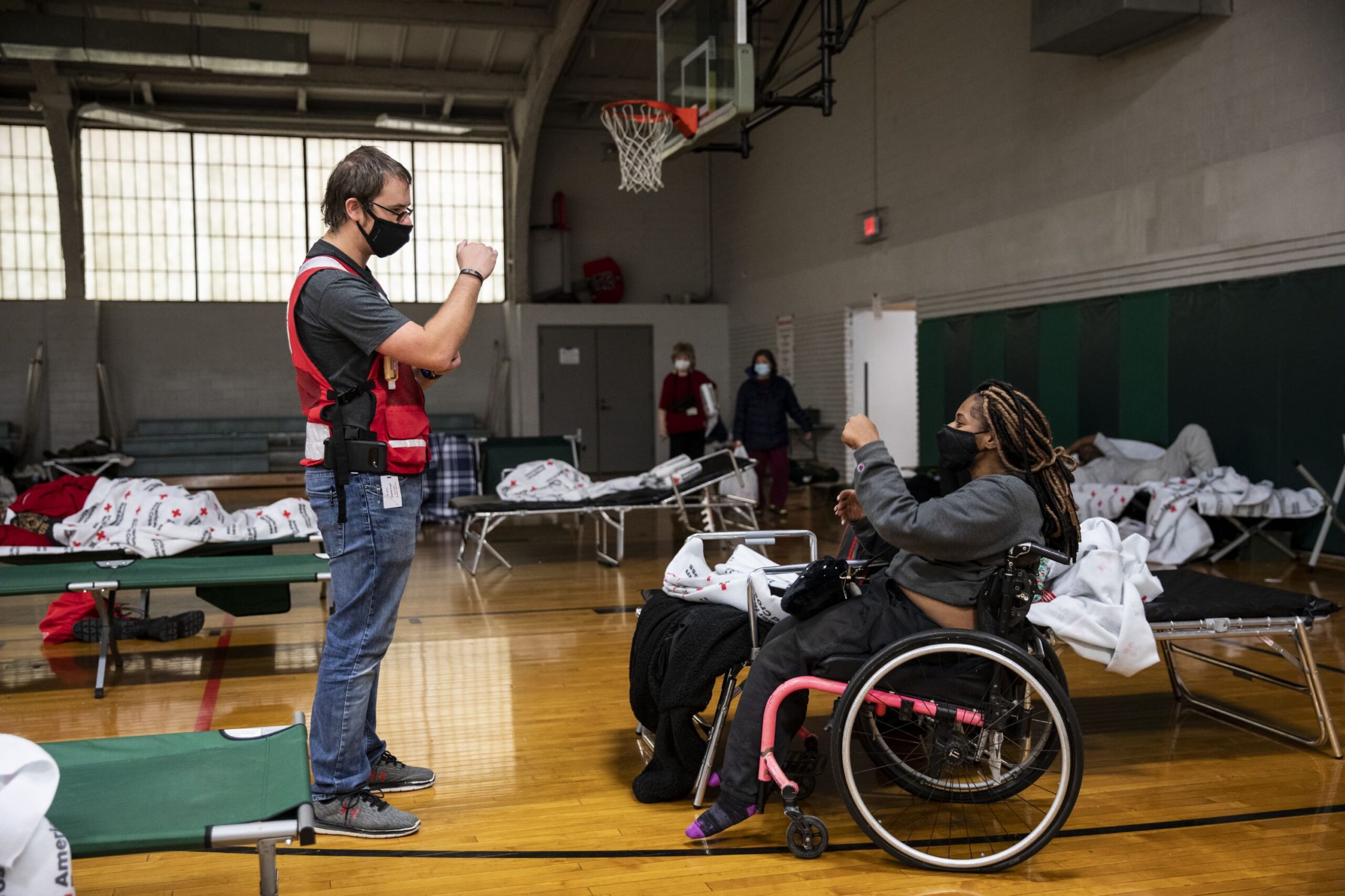 The image depicts disability inclusion during an emergency response situation, with an individual in a wheelchair being assisted by a Red Cross volunteer in a gymnasium temporarily converted into a shelter.