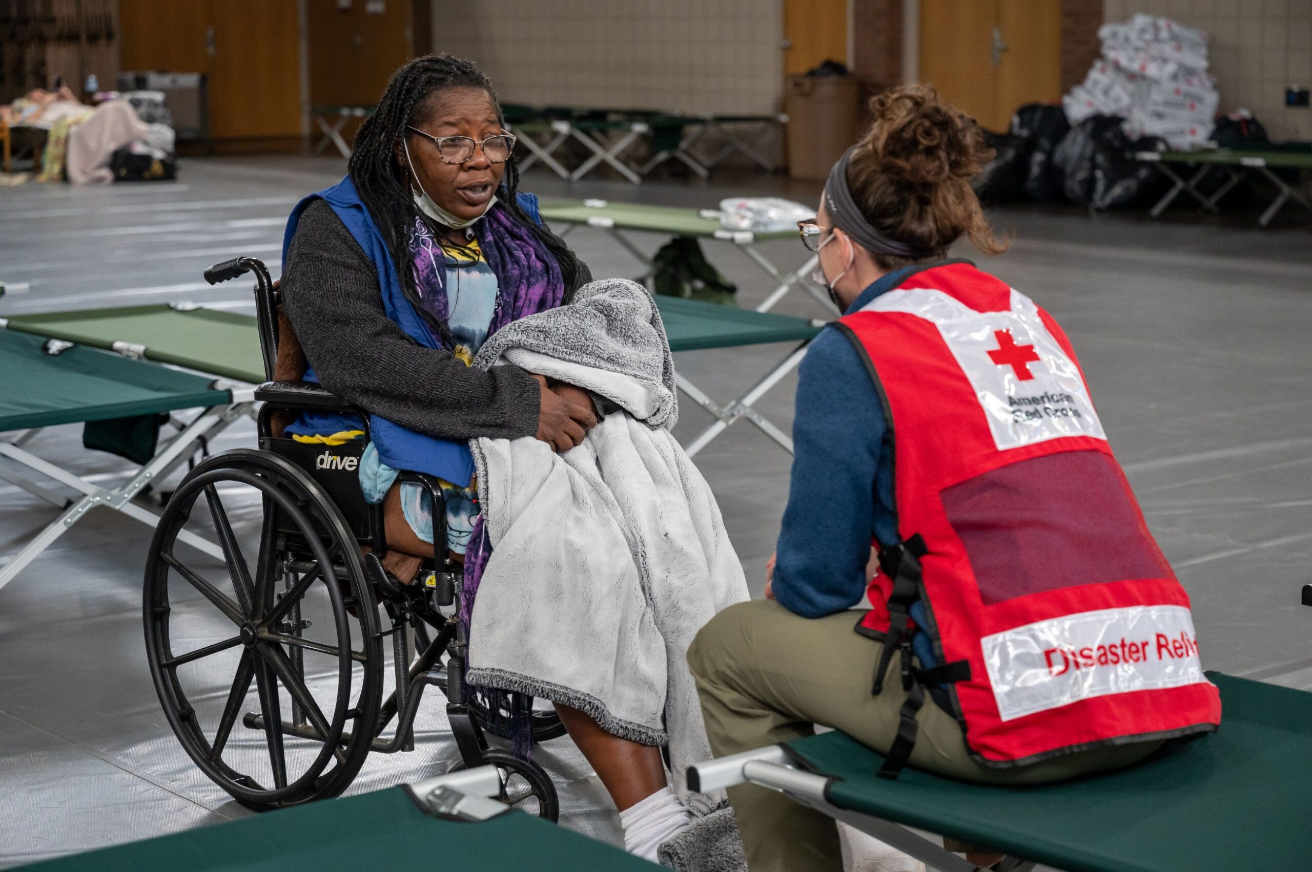 A disaster relief shelter, where a Red Cross worker wearing a vest is providing assistance to an elderly woman in a wheelchair.