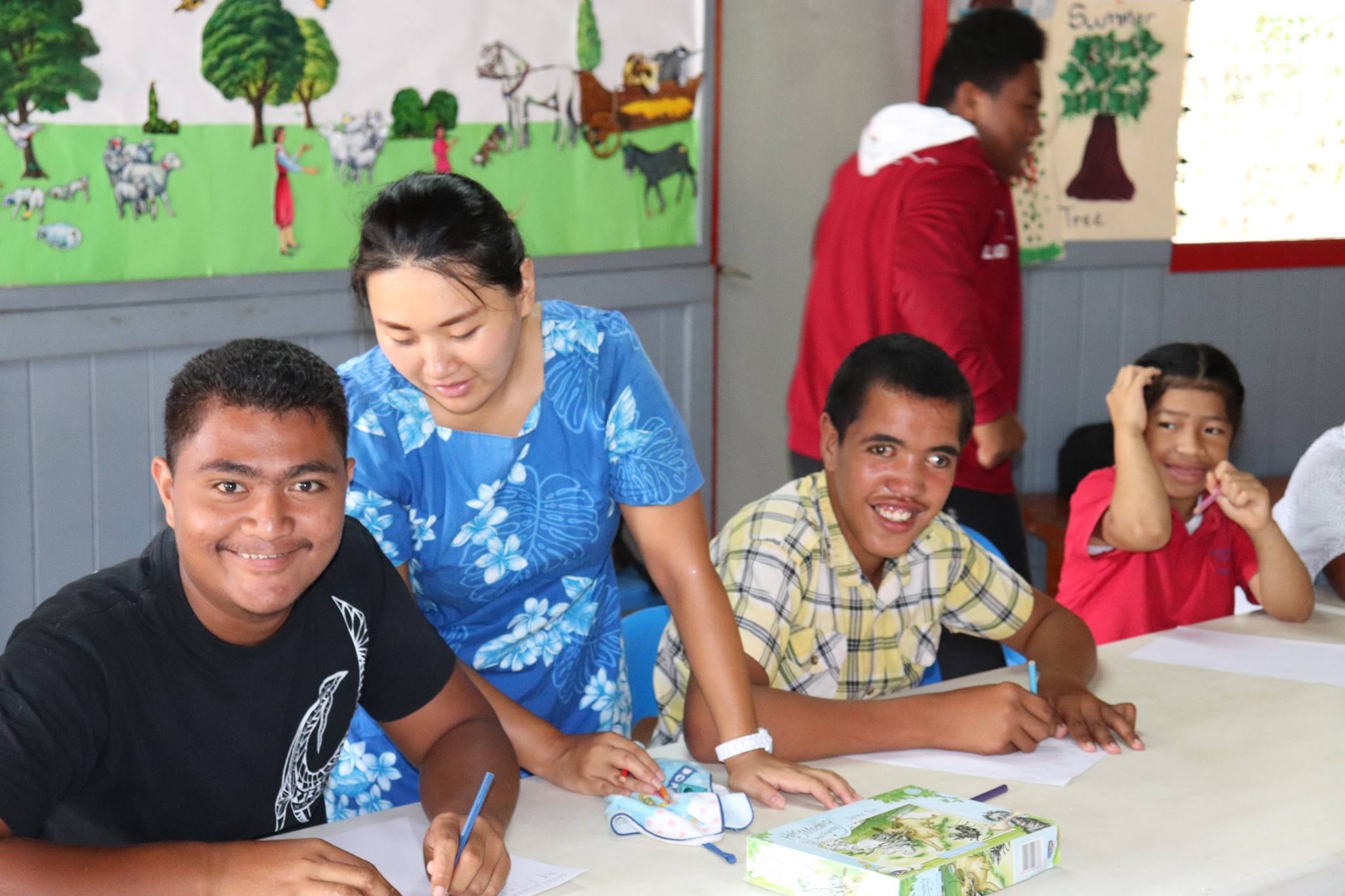A classroom with students with disabilities and a teacher engaged in learning activities. The teacher, wearing a blue floral dress, is assisting two male students sitting at a table and working on something together. The classroom environment appears inclusive and supportive for students with various abilities or needs.