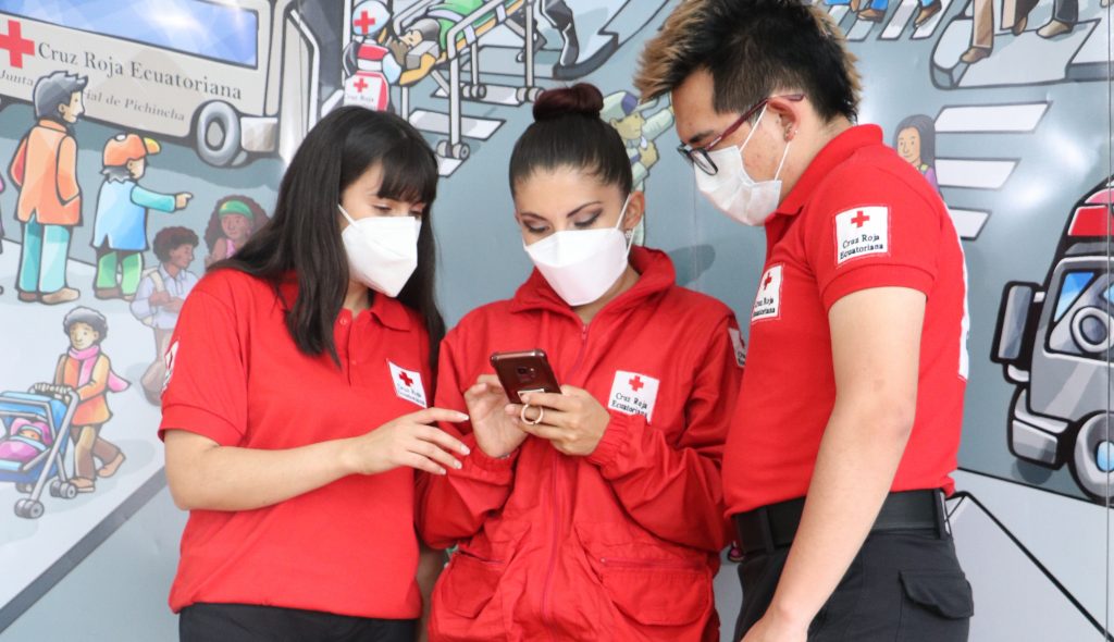 Three individuals wearing red Red Cross volunteer/staff shirts and face masks, gathered around a smartphone, seemingly coordinating efforts or accessing information in front of a mural related to humanitarian aid and assistance provided by the Ecuadorian Red Cross.