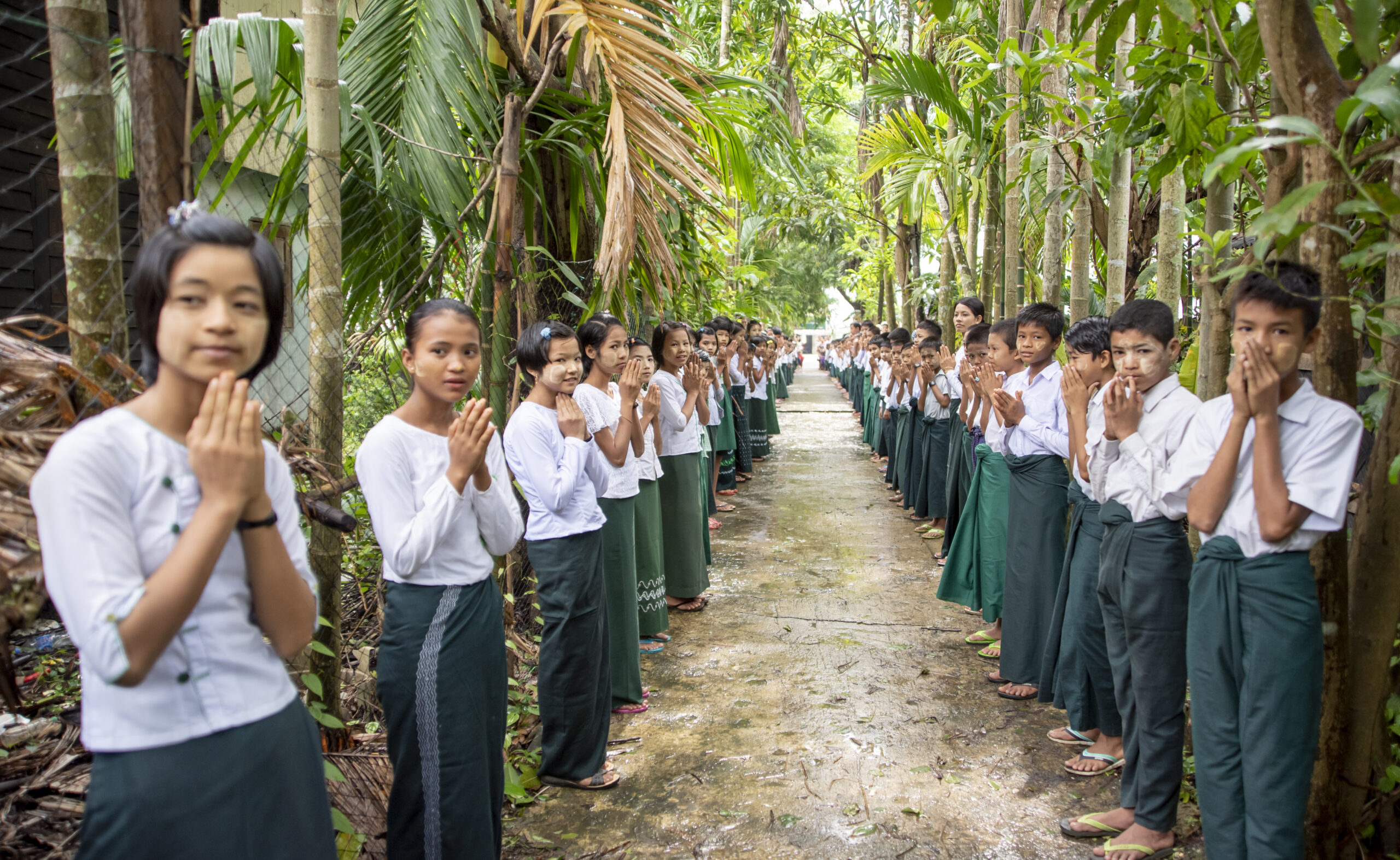 July 8, 2019. Sein Lay Kone village, Irrawaddy Delta, Myanmar. Children in a rural village in Myanmar welcome visitors with a traditional greeting. 
“The whole village is cooperating to teach the kids in this school about emergencies,” says Swe Swe Win–principal at the only school in Sein Lay Kone village, Myanmar where students (pictured) practice drop-and-cover drills to prepare for earthquakes. The community—located on a river close to the ocean—is only accessible by weaving through narrow, verdant channels by boat. Its residents are at-risk of cyclones, storms, floods, tsunamis, and earthquakes. Parents and non-parents alike have come together to ensure residents—especially kids—know what to do in case of crises. The Red Cross has stepped up to train students at the school on first aid and light search-and-rescue. “This training is not only useful at the school, but also so kids can treat their families and people in the community,” comments Swe Swe Win. The Red Cross has also distributed equipment such as solar panels, an early warning system, and first aid kits to the school.
The American Red Cross works alongside the Myanmar Red Cross to prepare disaster-prone communities for cyclones, floods, tsunamis, earthquakes, and other emergencies. We train and equip families with the tools they need to mitigate natural disaster risks and to be first responders when crises strike. In Myanmar, the American Red Cross teaches basic first aid, light search-and-rescue, and post-disaster epidemic control in 20 communities—in addition to running disaster simulations and forming village committees who mobilize when disasters hit. In 24 schools, we teach students basic first aid, light search-and-rescue, evacuation activities, and distribute emergency equipment—such as solar panels, fire extinguishers, megaphones, early warning speakers, first aid kits, and helmets. In Myanmar, some American Red Cross project sites are urban, while others sit in river deltas only accessible by