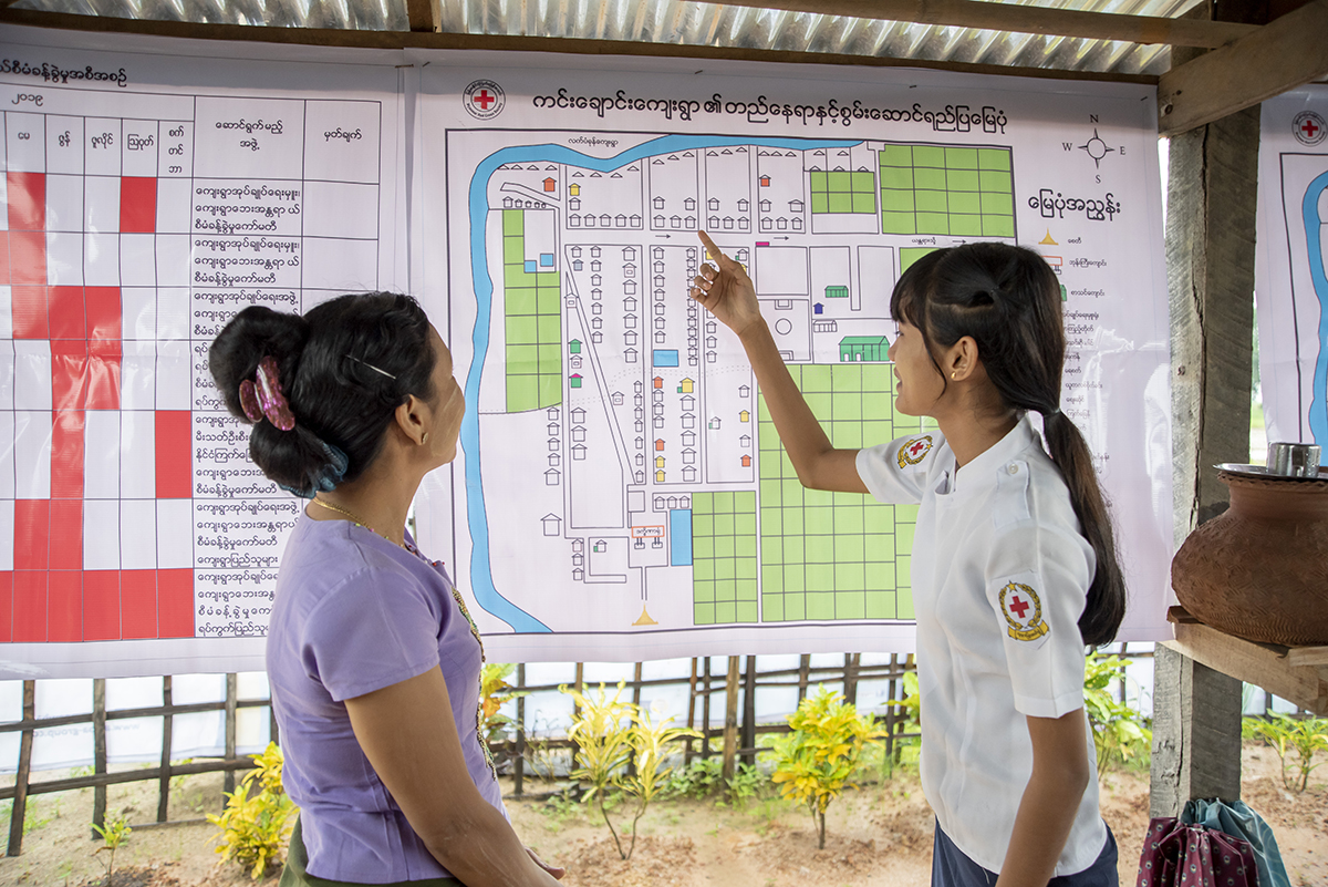 July 9, 2019. Kim Chaung village, Bago, Myanmar. Residents (Ma Sandar Win and Ma Han Thae Oo) of Kim Chaung village, Myanmar discuss a community map hanging in the town center. Developed with help from the Red Cross, the map can be used to mitigate disaster risks. Communities like this one work together to identify their risks—such as flood zones— as well as resources they can utilize during disasters. Maps show homes, high-risk areas, evacuation routes, locations of handicapped residents and the elderly (who may need help evacuating), and safe spaces to seek shelter. This rural community—dotted with rice patties and farmland—experiences regular floods and is at-risk of earthquakes and strong storms.

The American Red Cross works alongside the Myanmar Red Cross to prepare disaster-prone communities for cyclones, floods, tsunamis, earthquakes, and other emergencies. We train and equip families with the tools they need to mitigate natural disaster risks and to be first responders when crises strike. In Myanmar, the American Red Cross teaches basic first aid, light search-and-rescue, and post-disaster epidemic control in 20 communities—in addition to running disaster simulations and forming village committees who mobilize when disasters hit. In 24 schools, we teach students basic first aid, light search-and-rescue, evacuation activities, and distribute emergency equipment—such as solar panels, fire extinguishers, megaphones, early warning speakers, first aid kits, and helmets. In Myanmar, some American Red Cross project sites are urban, while others sit in river deltas only accessible by boat. Note: The country of Myanmar is also known as “Burma.” Photo by Brad Zerivitz/American Red Cross