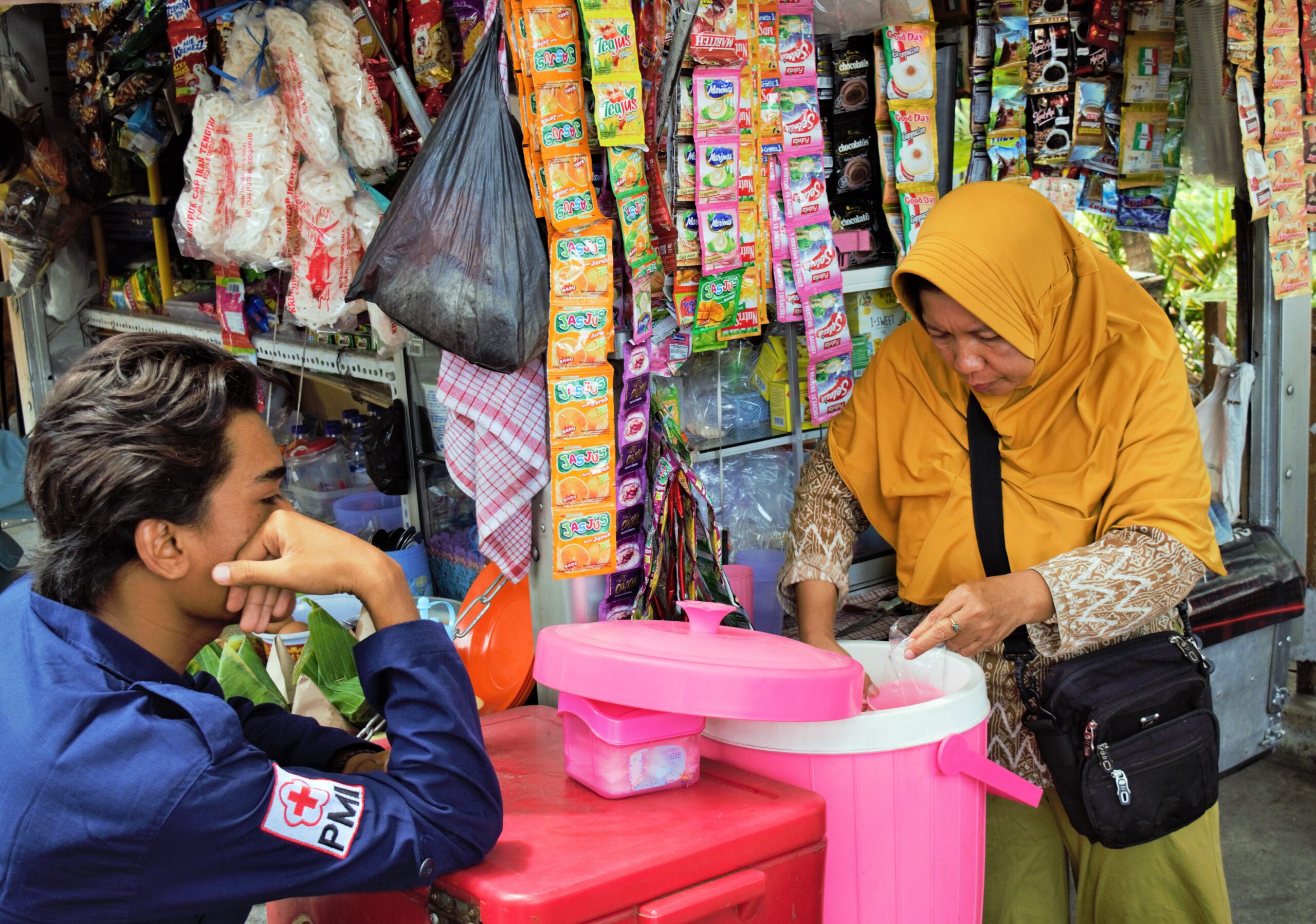 A female shopkeeper in Indonesia scooping water from a pink bucket, serving a Red Cross Volunteer in a blue shirt.