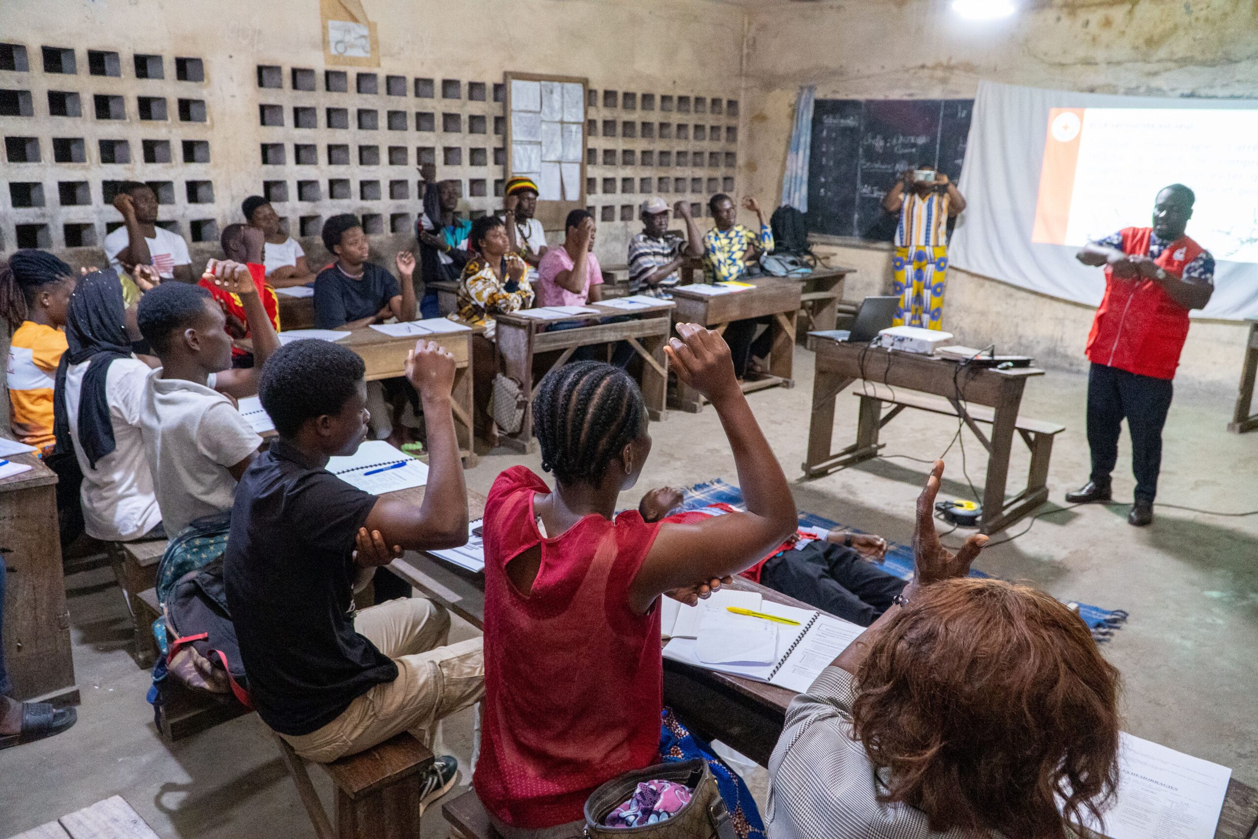A group of people sitting at desks in a classroom with their hands raised. A Red Cross volunteer stands at the front teaching them first aid skills.