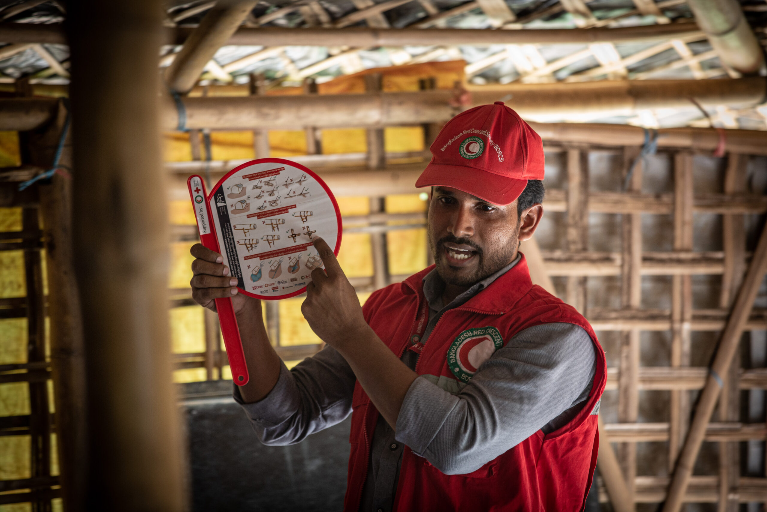 A Red Crescent volunteer holds a sign with illustrated instructions demonstrating how to secure shelters ahead of monsoon rains or cyclones.