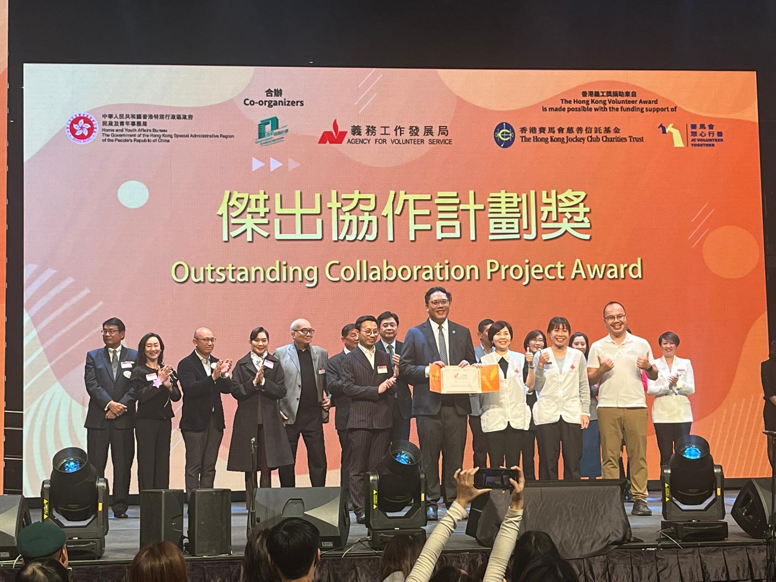 A big group of people, including Hong Kong Red Cross staff, holding an award certificate on stage. A screen in the background says Outstanding Collaboration Project Award.