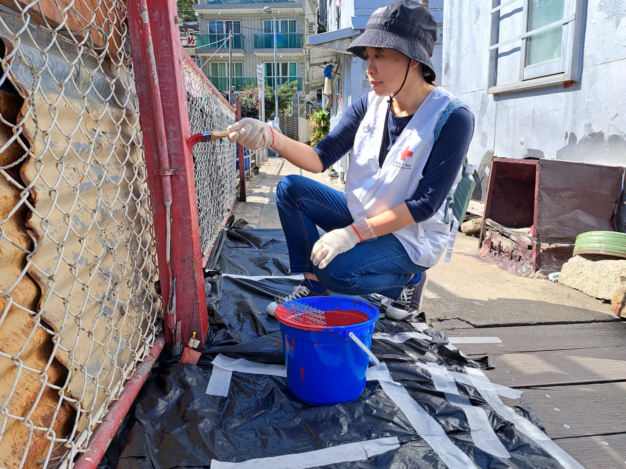 Eva Yeung, wearing a white vest with the Hong Kong Red Cross logo, is seated and crouching down, painting the metal poles of a welded wire fence red.