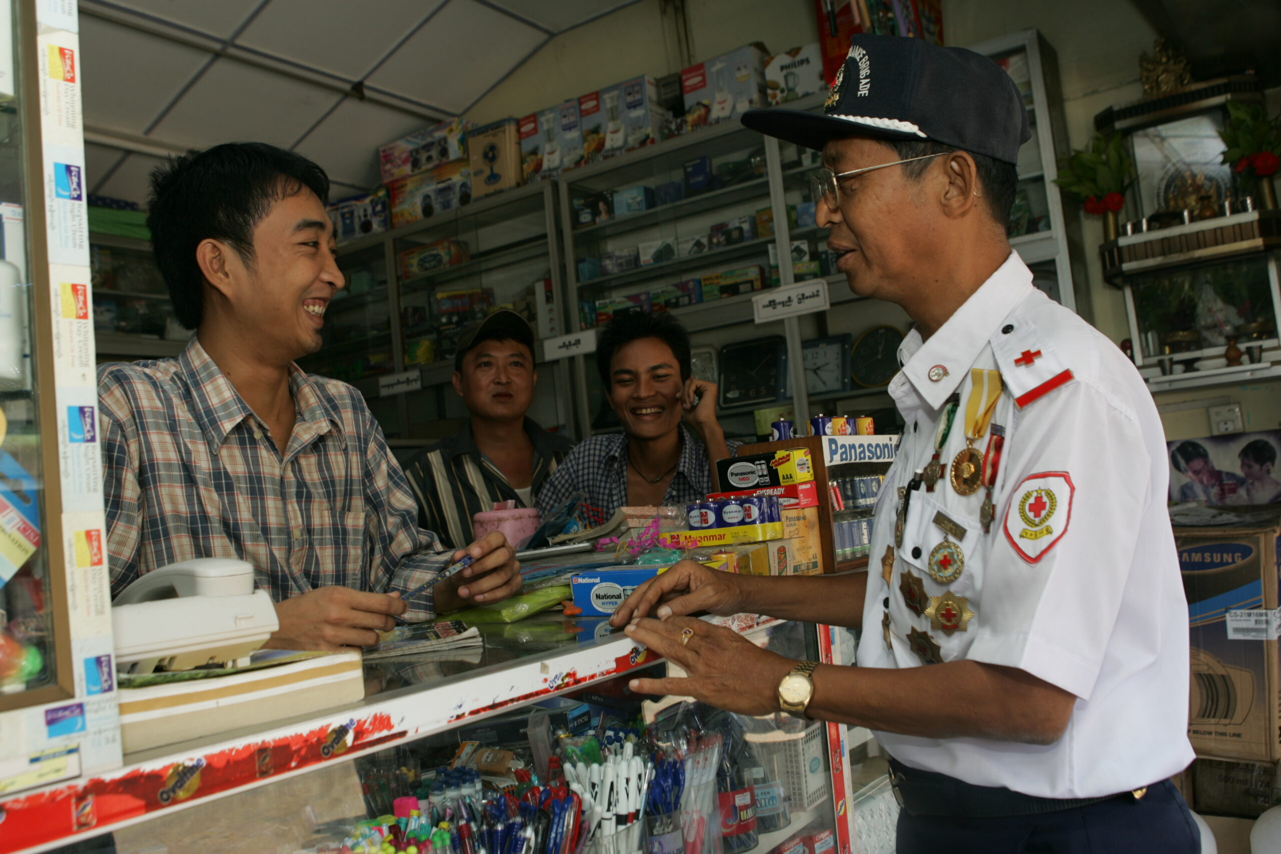 Maubin Township Red Cross is the strongest branch of Myanmar Red Cross Society. It has a monthly income of more than 300,000 kyats (about 300 US dollars) while most branches can hardly make 20-30,000 kyats. It has got bottled water business, inter-city buses licensed under the branch, and rented out a few stalls at the main market. It has its own office and rents part of its office for Desktop Publishing for income generation.
Because it is located in Ayeyarwady delta, volunteers from the branch sometimes help out in times of boat mishaps or ferry disasters. Quite a few volunteers are trained in water safety or lifeguard training.
U Myint Wai, staff officer grade 2 of Maubin district Red Cross, talks with the owner of a stall rented out by the red cross.