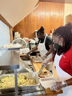 CAP Team support local nonpprofits Dr. April Masha Jones and a team member wearing gloves and aprons prepare takeout food containers in a kitchen, standing behind trays of cooked food.