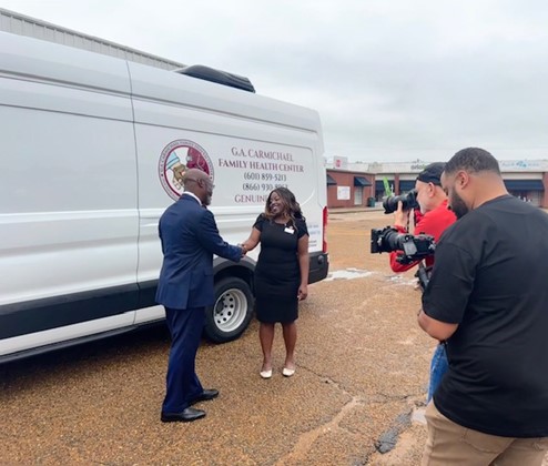 Carmichael Family Health Center Dr. April Masha Jones and Dr. James Coleman shake hands in front of a G.A. Carmichael Family Health Center mobile medical van, while a film crew records the moment.