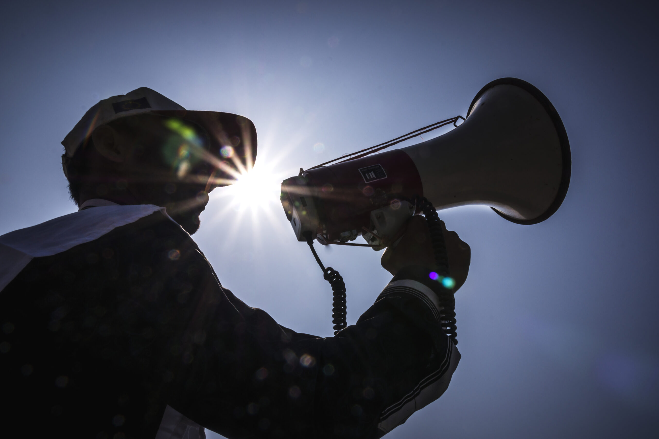 A silhouetted community volunteer uses a megaphone to deliver an early warning message outdoors, with sunlight behind them against a clear sky.