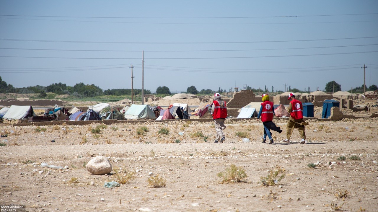 Afghan Red Crescent Society volunteers in red vests walk across a dry, arid landscape towards a temporary displacement camp with rows of tents in Afghanistan.