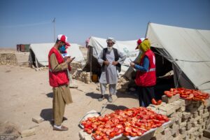 Two Afghan Red Crescent Society volunteers conduct a survey with a community member outside a tent at a displacement site in Balkh Province, northern Afghanistan, with tomatoes drying in the foreground.