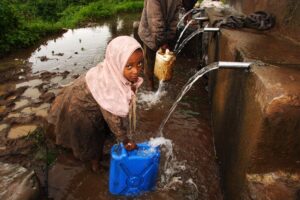 A young girl in a pink hijab collects water from a tap into a blue jerrycan in muddy conditions in Ethiopia.