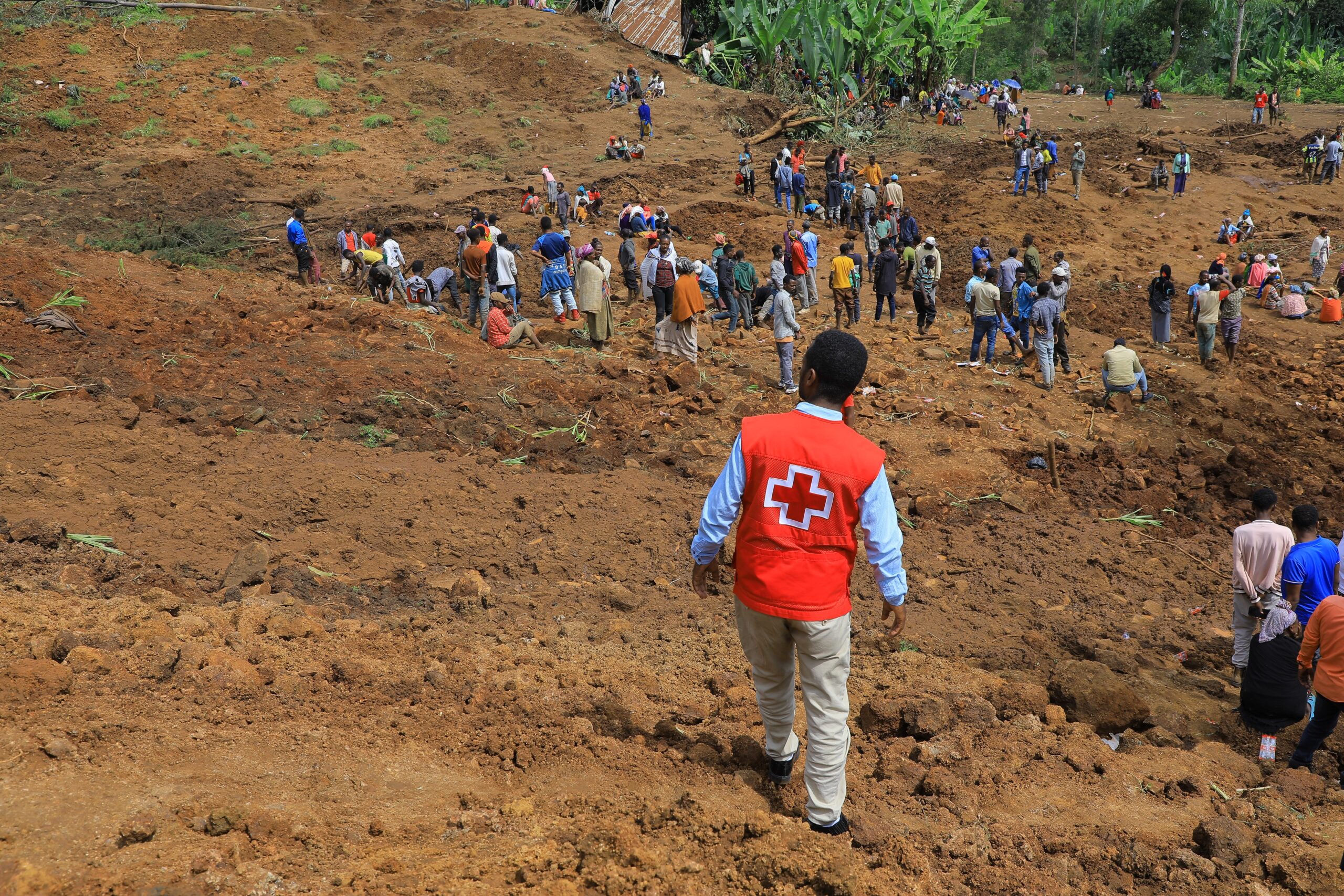 An Ethiopian Red Cross volunteer in an orange vest walks towards a large crowd of people gathered at the site of a landslide in Ethiopia.