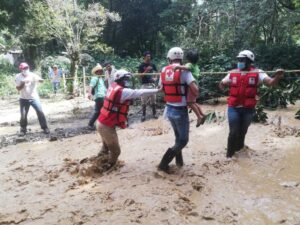 Red Cross volunteers in helmets and life jackets carry a child across a fast-moving muddy river using a rope line during a flood rescue operation in Central America.