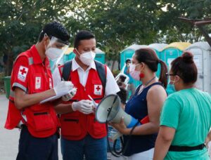 wo Honduran Red Cross volunteers wearing masks and face shields take notes on a clipboard while speaking with community members holding a megaphone at an evacuation site in Honduras.