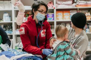 A Hungarian Red Cross volunteer wearing a mask and stethoscope examines a young child held by their caregiver at a relief distribution point in Hungary.