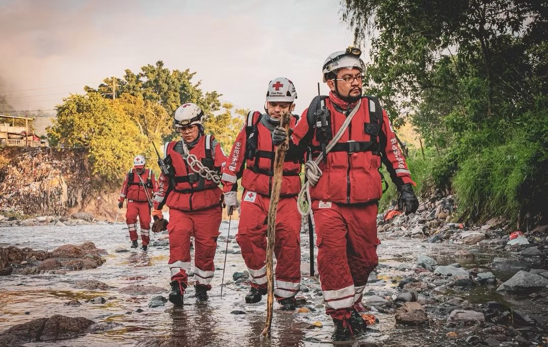 A team of Guatemalan Red Cross rescue volunteers in full red gear, helmets and life jackets walk along a rocky riverbed carrying ropes and equipment during an emergency response operation.