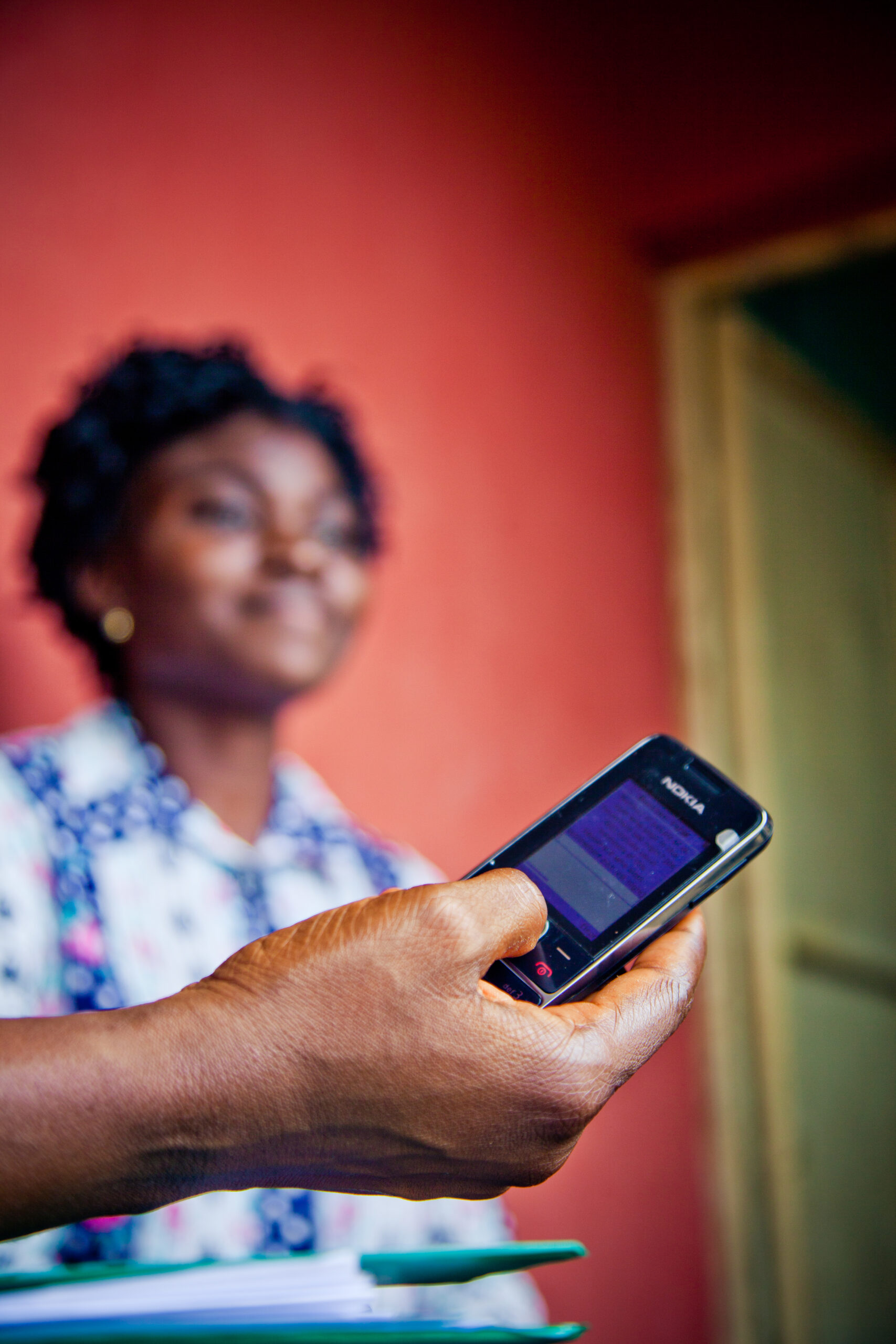 A hand holds a cellphone running a survey app. A woman, out of focus, sits in the background against a red wall.