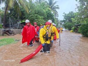 Salvadorian Red Cross volunteers in helmets and rain gear wade through deep floodwaters carrying a rescue board during a flood response operation in El Salvador.