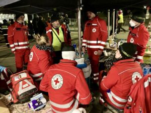 A group of Slovak Red Cross volunteers in uniform gather for a briefing at an outdoor reception point for Ukrainian refugees at the Slovak border at night.