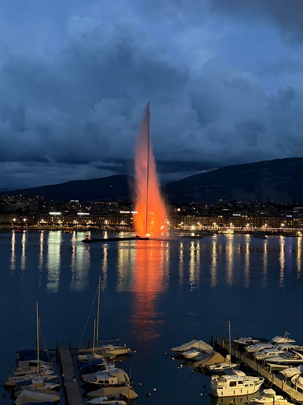 A tall fountain jet lit bright orange rises over Lake Geneva at twilight, reflected in the water beside a marina of moored boats, with the city skyline and hills behind.