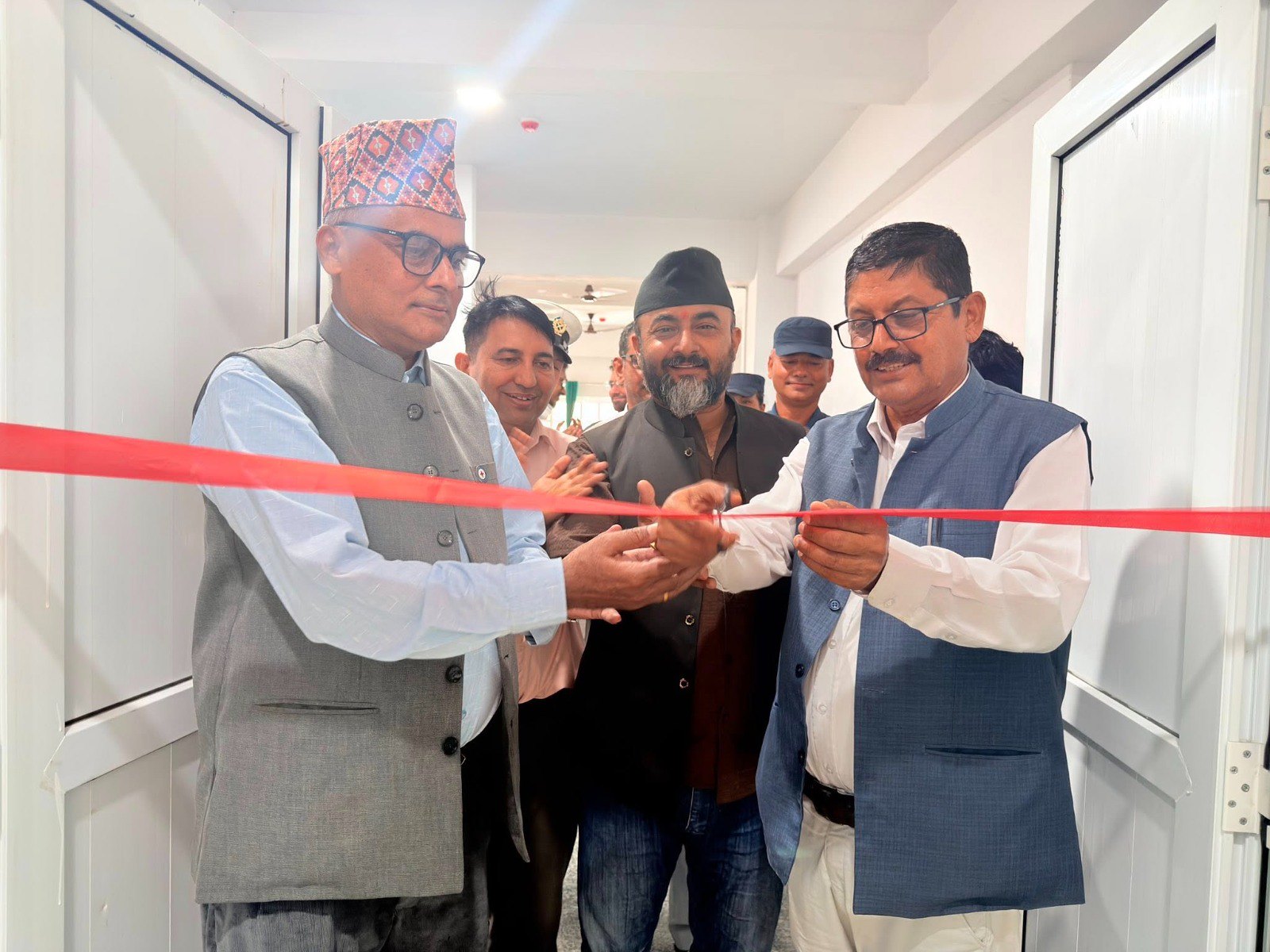 Several men in formal vests, two wearing traditional Nepali caps, cut a red ribbon together in a bright hospital corridor as onlookers smile.