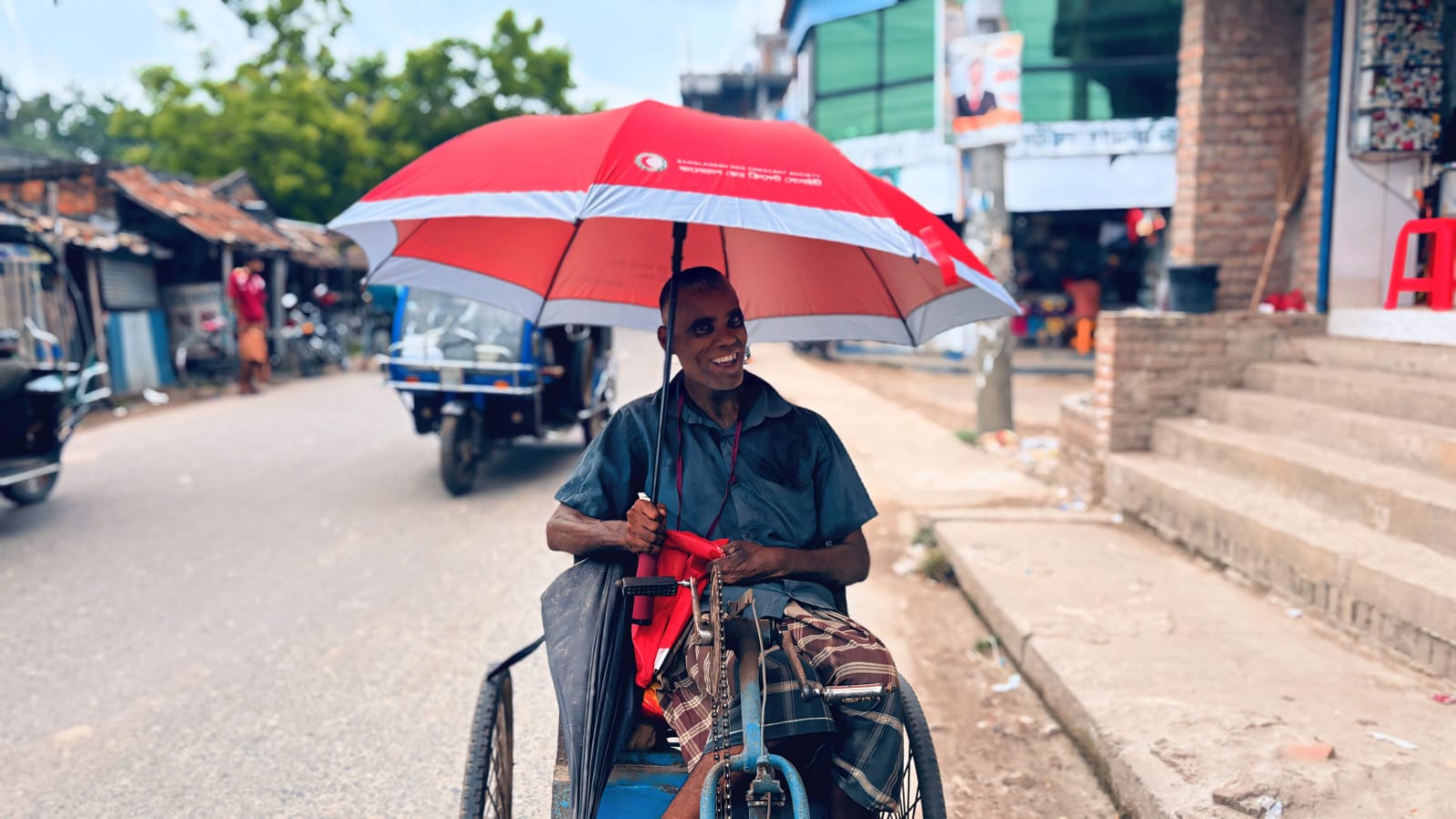 A man seated on a hand-pedaled tricycle on a sunny street shelters under a large red-and-white Red Crescent umbrella, smiling toward the camera.