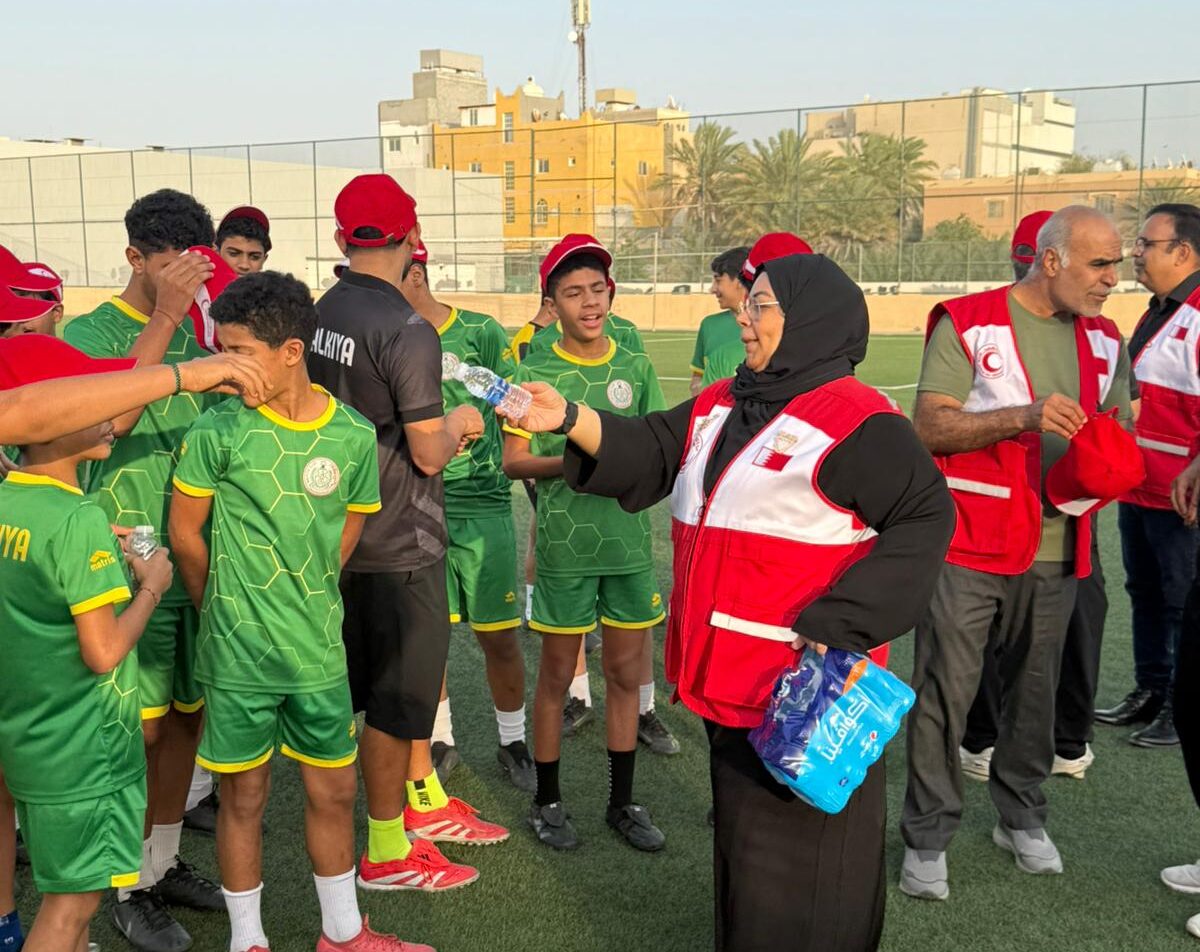 Red Crescent volunteers in red vests speak with a group of children in green football kits and red caps on a turf pitch, some drinking water, with buildings and palm trees behind.