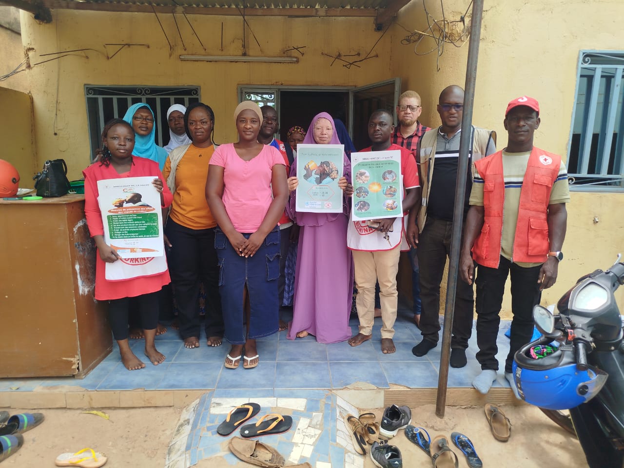 A group of people, including Burkina Faso Red Cross volunteers in red vests and Ministry of Health staff, stand together outside a building holding printed heat-awareness posters.