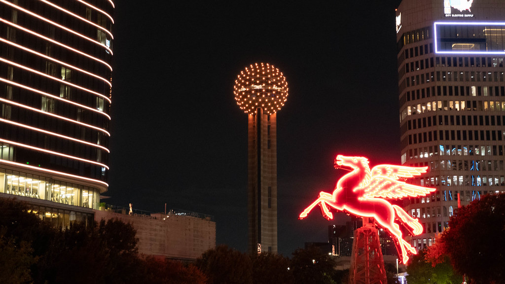 The illuminated spherical top of Reunion Tower and a glowing red neon Pegasus sign rise above the Dallas skyline against a dark night sky.
