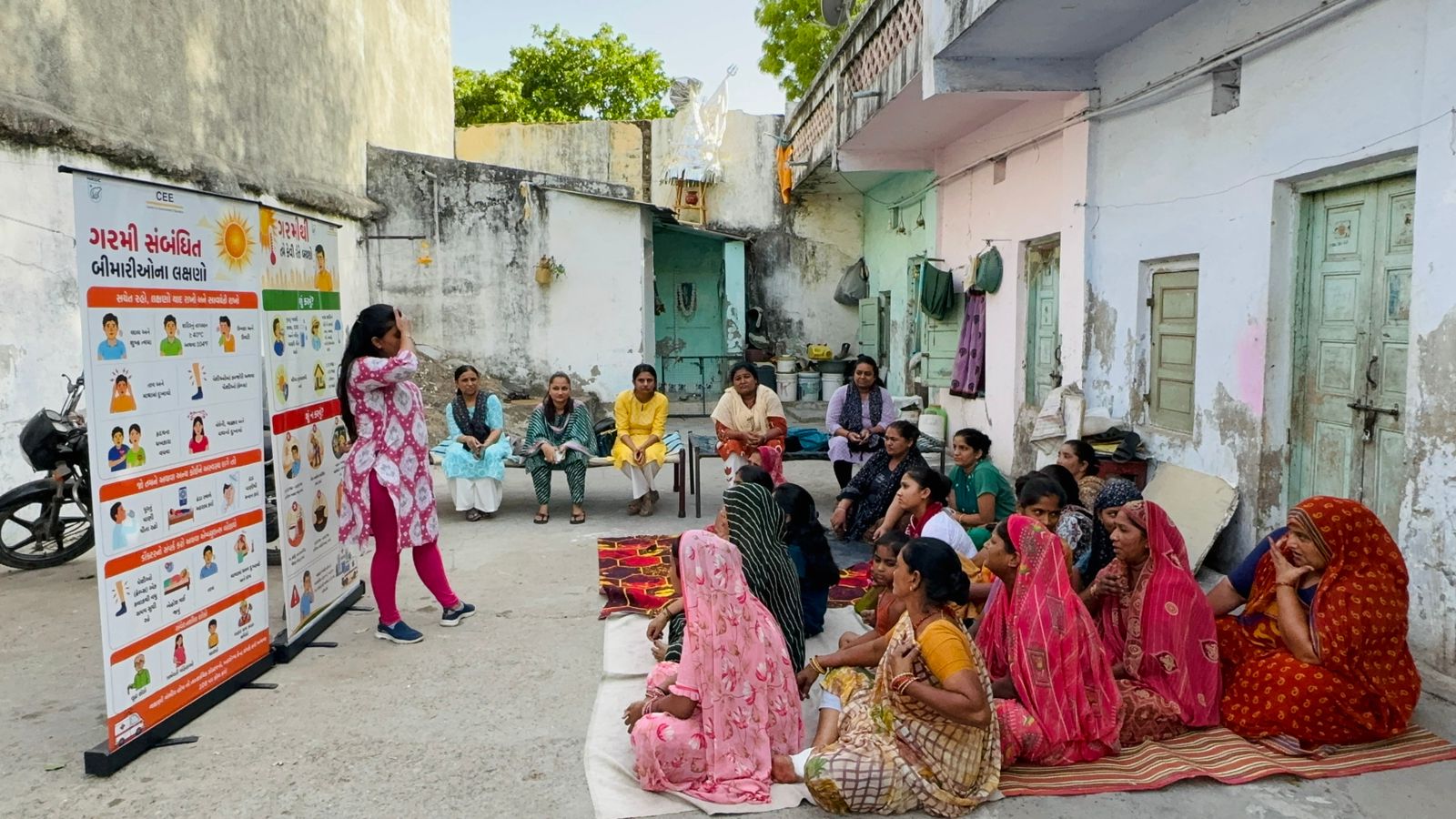 A woman stands beside illustrated heat-illness posters addressing a seated group of women gathered on mats in an open courtyard.