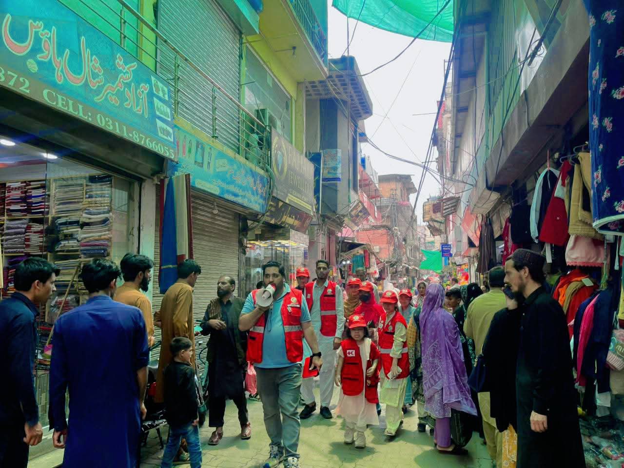 A volunteer in a red vest speaks into a megaphone while walking through a narrow, busy market street lined with shops, accompanied by other volunteers including children in matching vests.