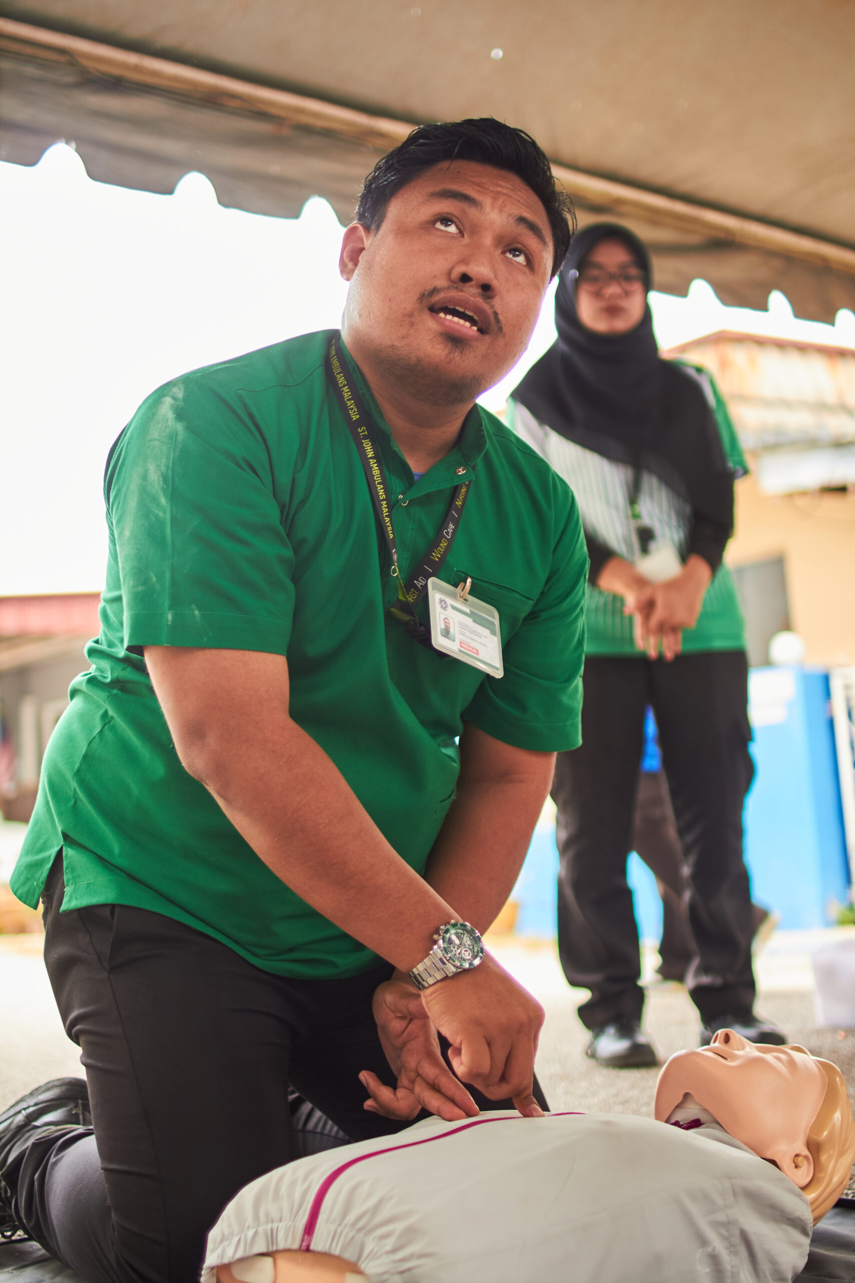 A first-aid instructor in a green shirt kneels beside a training manikin, speaking to participants under a shelter, with a colleague observing in the background.