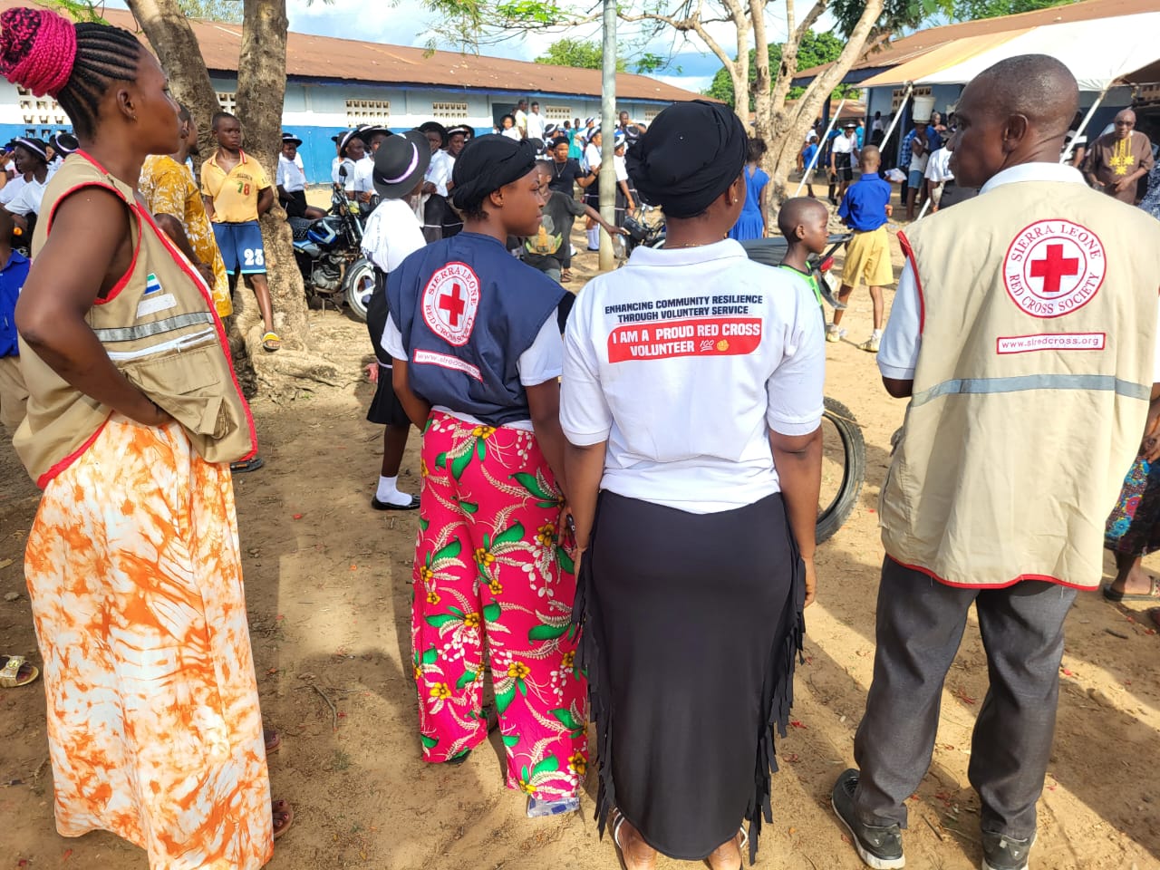 Sierra Leone Red Cross volunteers in branded vests stand with students outdoors on a school grounds, with a crowd of uniformed pupils gathered under trees in the background.