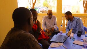 Three men, one wearing an SRCS red vest, huddle around laptops at a table in a sunlit room, collaborating on work during a training or planning session.