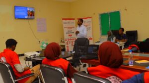 An SRCS trainer stands and presents to a small group of participants in red vests inside a meeting room at the SRCS Dhusamareb Branch, with a staff structure roll-up banner and a slide on flood response objectives displayed on a wall-mounted screen.