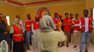 Somali Red Crescent Society (SRCS) staff and volunteers in red vests gather in a circle indoors for a briefing or training session, with the SRCS logo banner visible on the wall behind them.