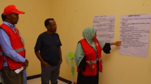 An SRCS volunteer in a red vest presents group work to two colleagues, pointing to flip chart papers on the wall showing a communication plan and a list of anticipatory actions for flood preparedness, including early warning, evacuation, pre-positioning of relief supplies, and health and WASH preparedness.