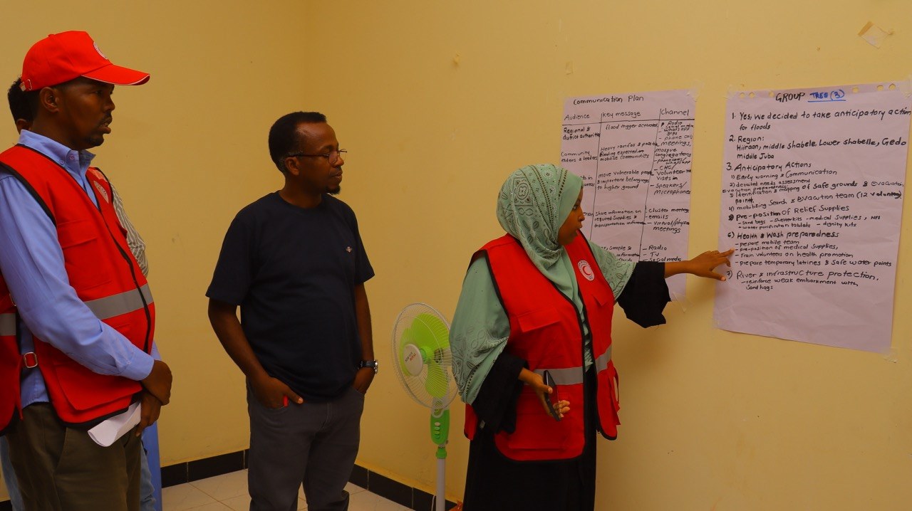 Three Somali Red Crescent participants, two in red vests, review handwritten flip-chart notes on a wall during the Dhusamareeb preparedness exercise.