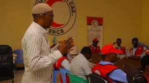 A man addresses a group of SRCS staff and volunteers seated around a table in a meeting room, with the SRCS logo on the wall behind him, during what appears to be a training or planning session.