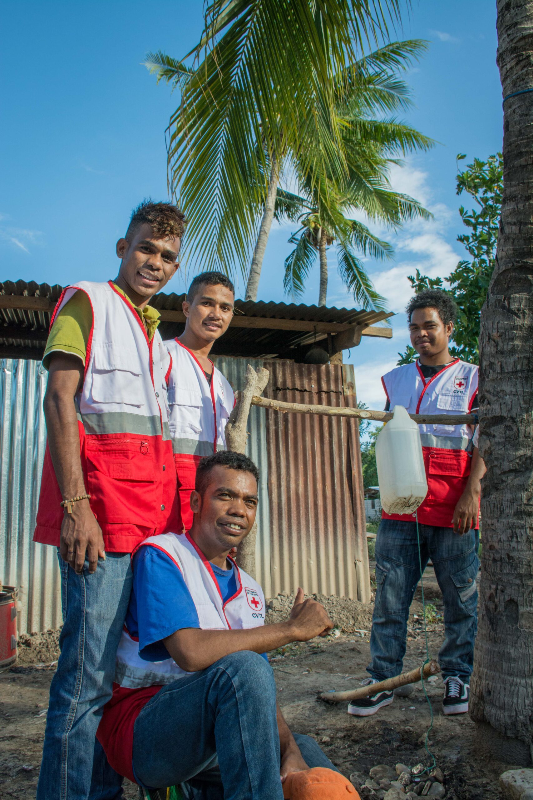 Four Timor-Leste Red Cross volunteers in red Red Cross vests stand and kneel together outdoors in a village, with palm trees and a corrugated-metal shelter behind them.