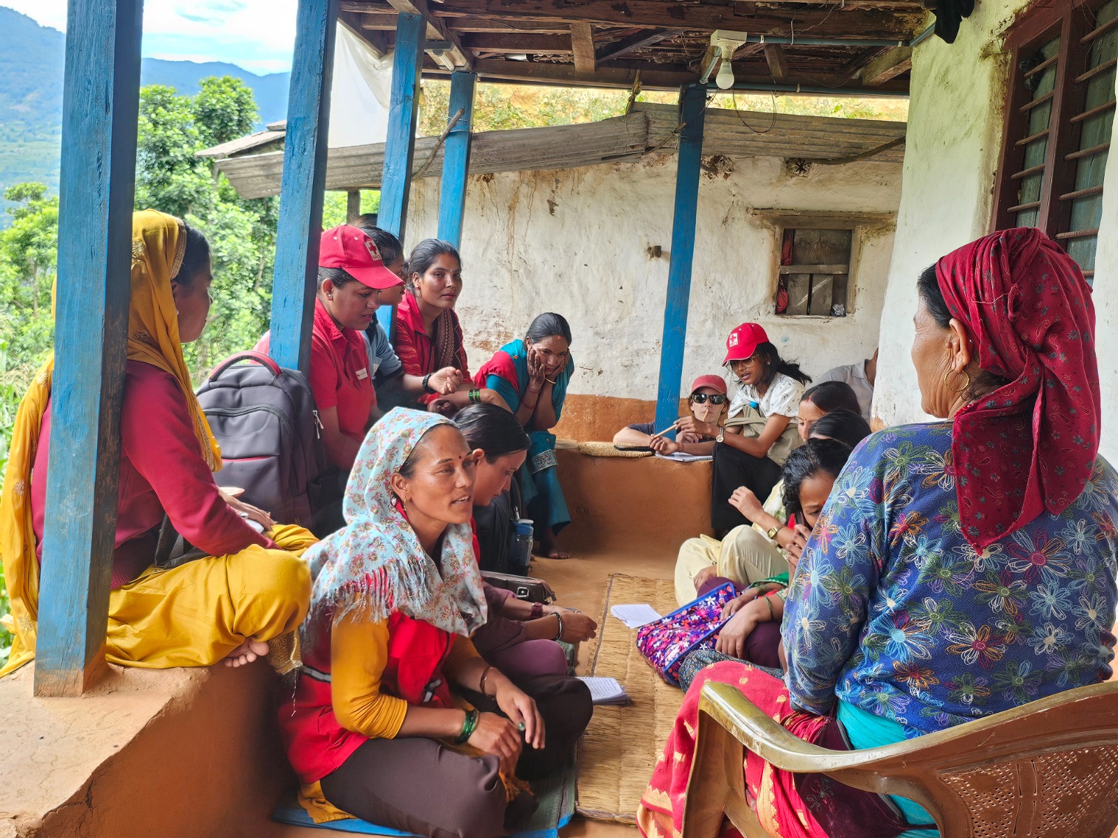 Red Cross volunteers and women from the community, some wearing headscarves and red caps, gather on a rural home's covered porch for a family-level discussion, with mountains visible in the background.