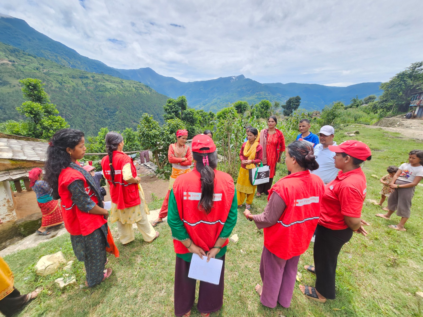 Nepal Red Cross Society volunteers in red vests and caps stand in a circle with community members on a hillside, discussing disaster risk reduction (DRR) against a backdrop of green mountains.